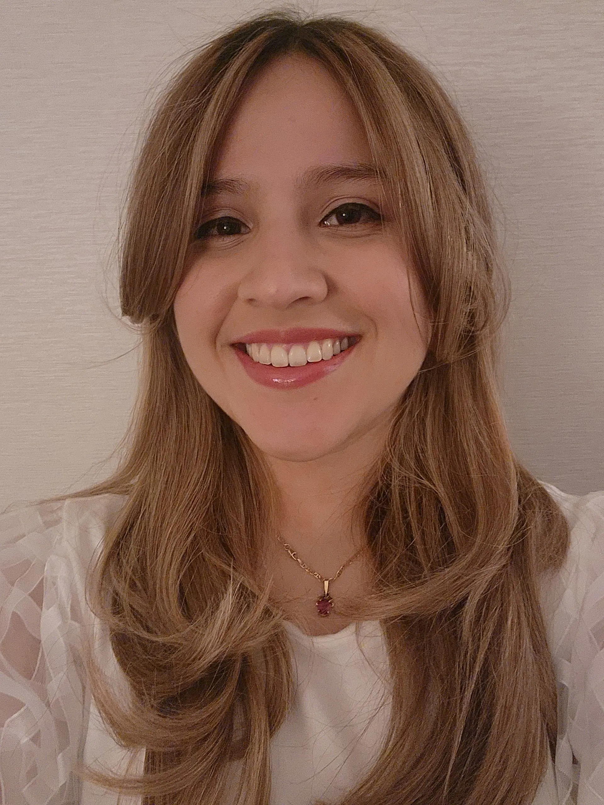 A woman smiles in a white shirt with heavily layered dark blonde hair parted down the middle