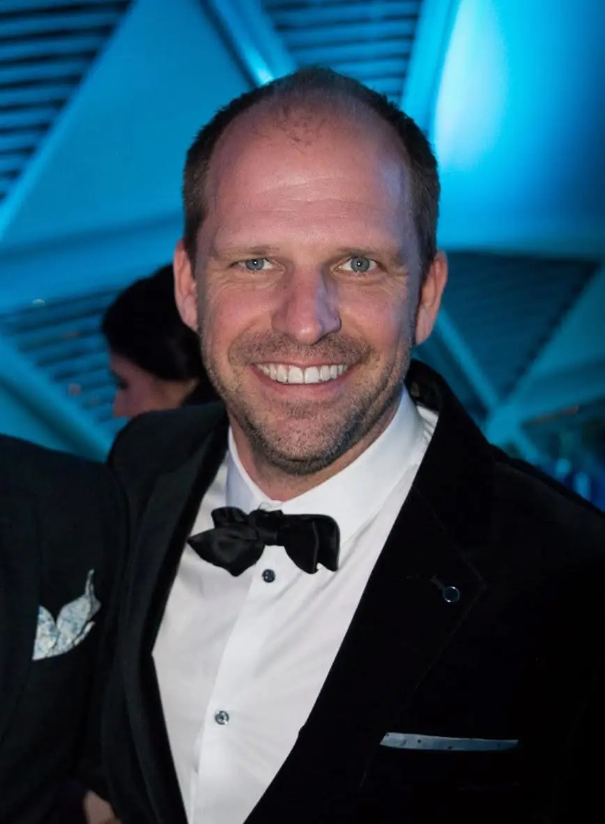 White man smiles in a black tux and bowtie with a blue background