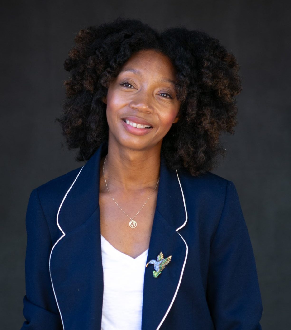 A woman with curly hair smiles while wearing a dark blazer with a hummingbird brooch